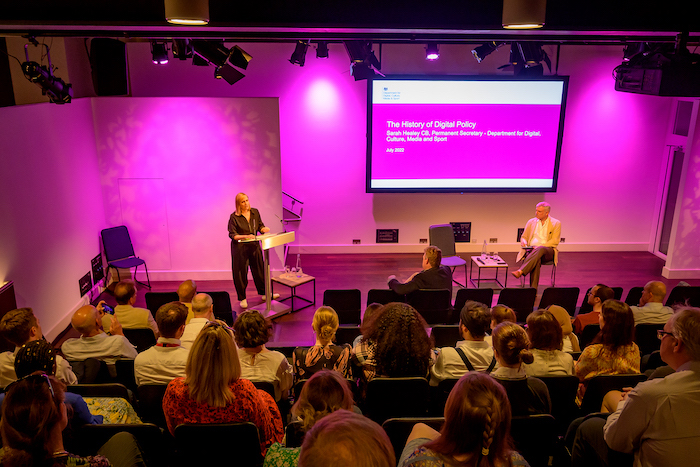 Sarah Healey, standing on a stage with a pink backdrop, in front of a seated audience 